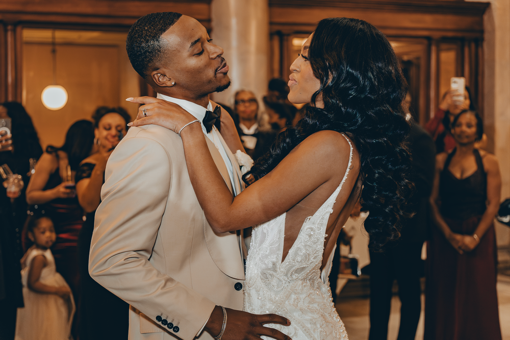 A couple shares a romantic first dance at their wedding reception while guests in black dresses watch in the background.