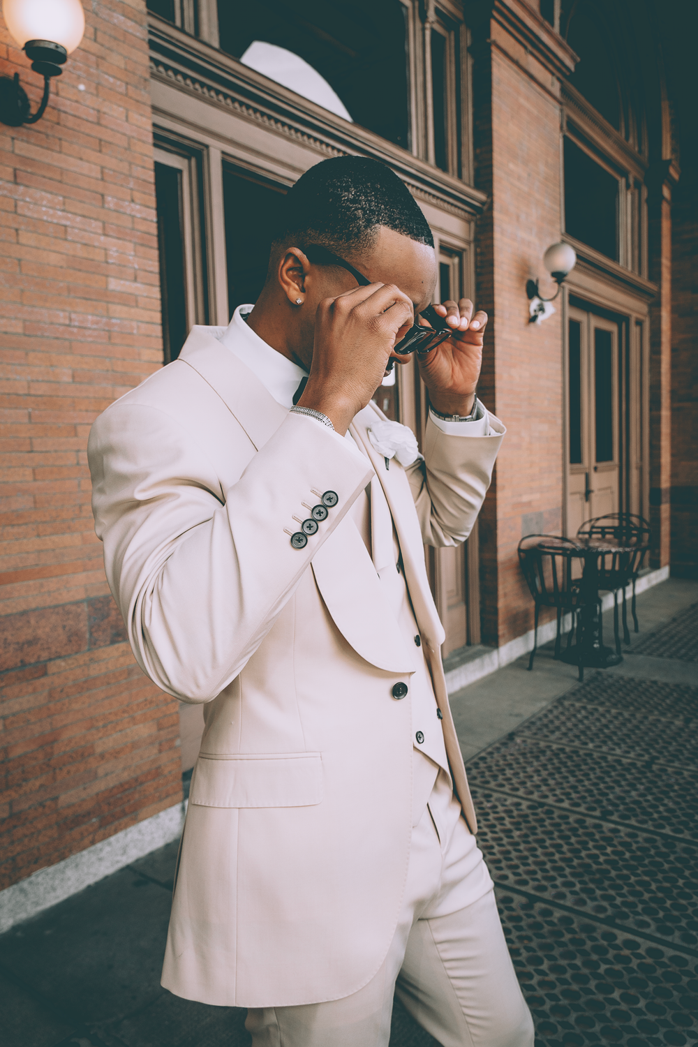 Person in cream colored suit touching their face while standing outside brick building on city street.