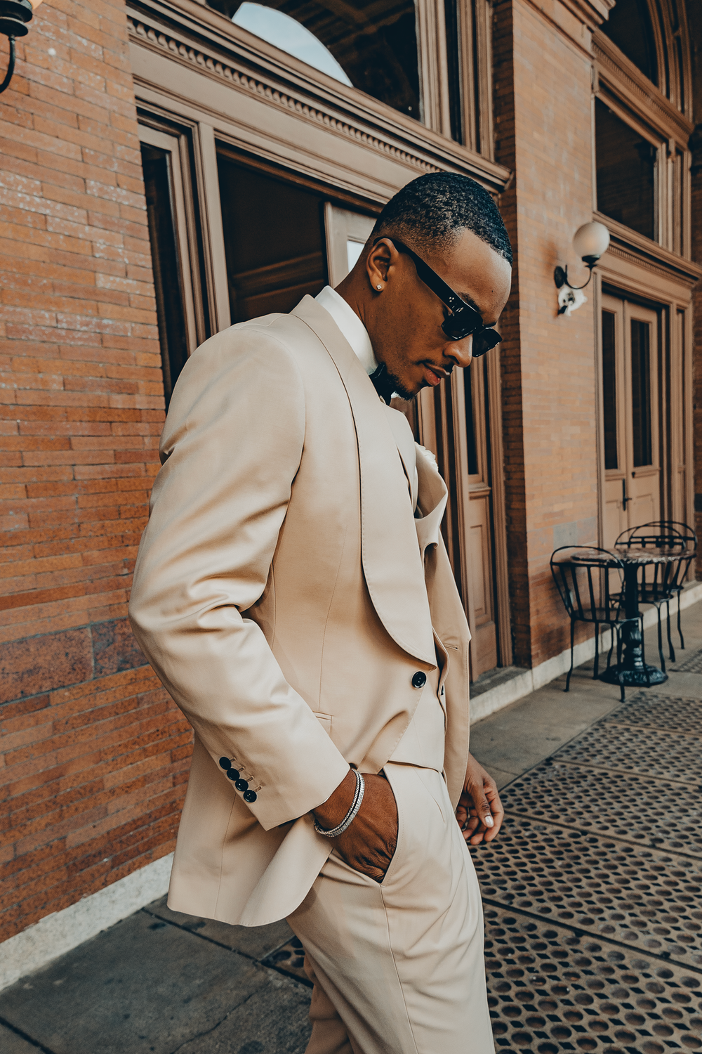 A man wearing a stylish cream-colored suit and sunglasses stands confidently against a brick building exterior.