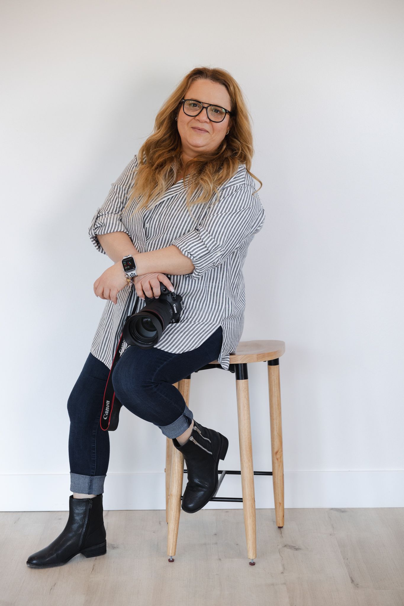 Person in striped shirt and rain boots poses on wooden stool with camera against white wall.
