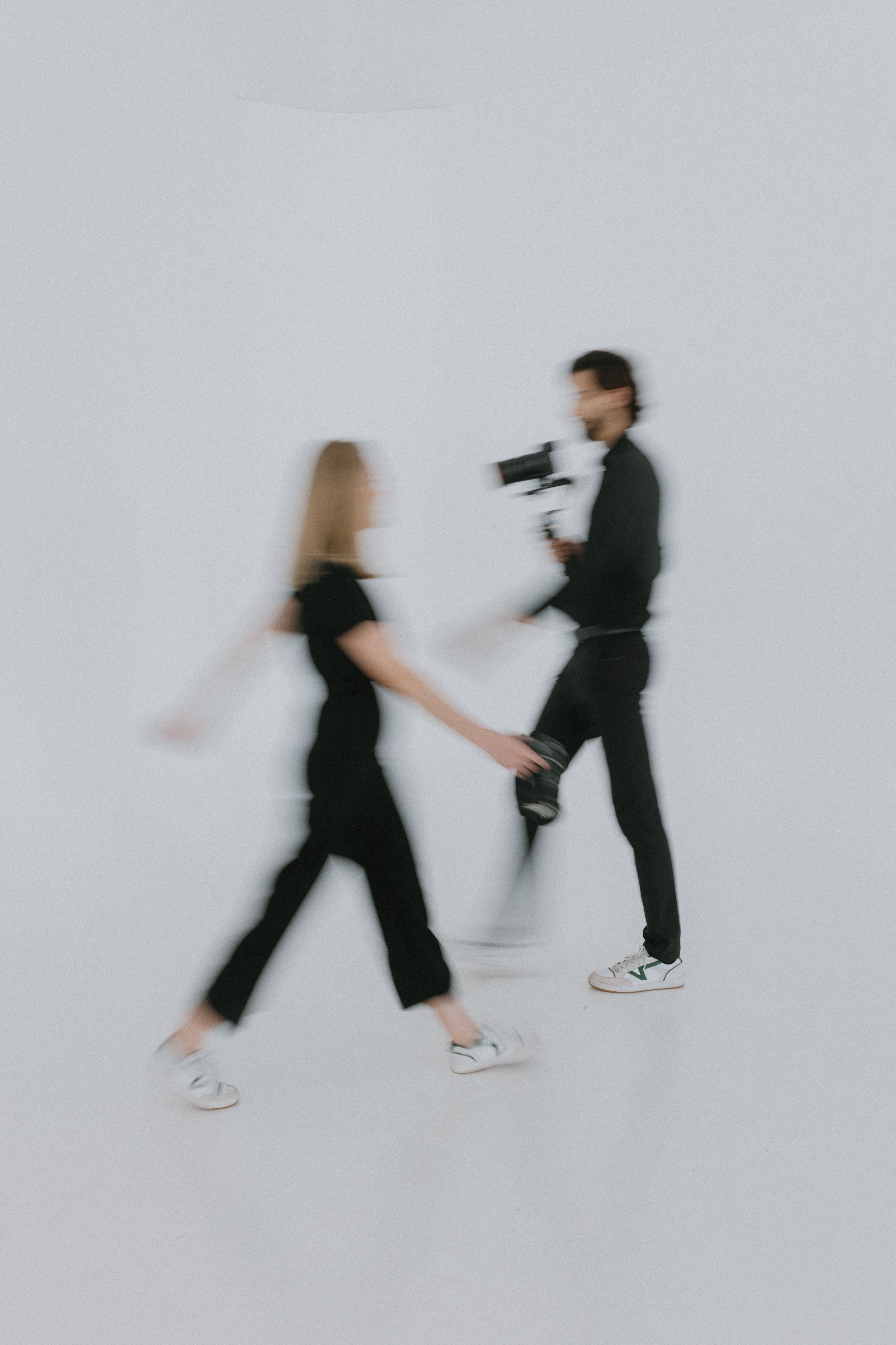 Motion blurred portrait of two people in black outfits walking past each other against a white backdrop.