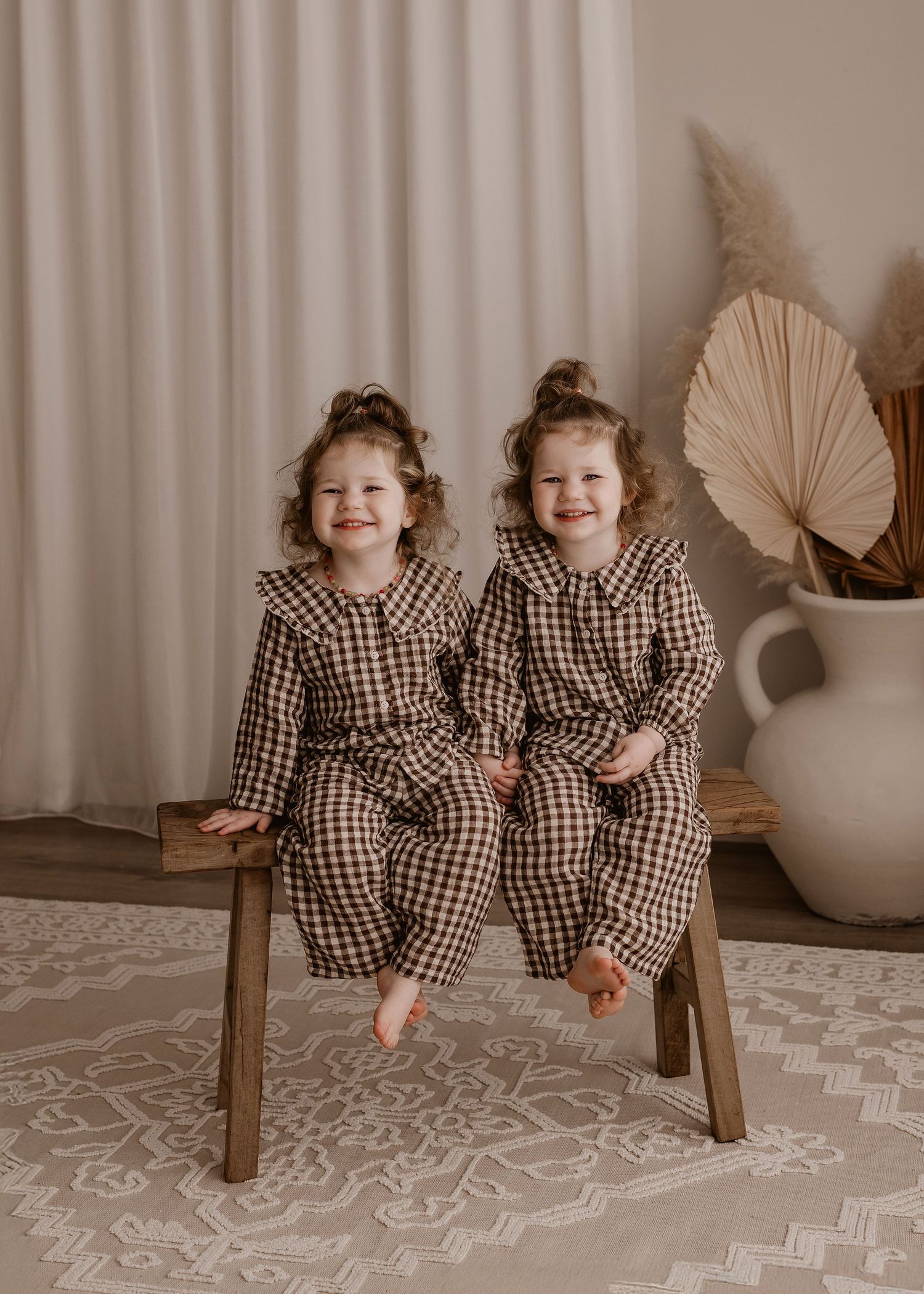 Two small children sitting together in matching beige outfits on wooden chair with rustic decor.