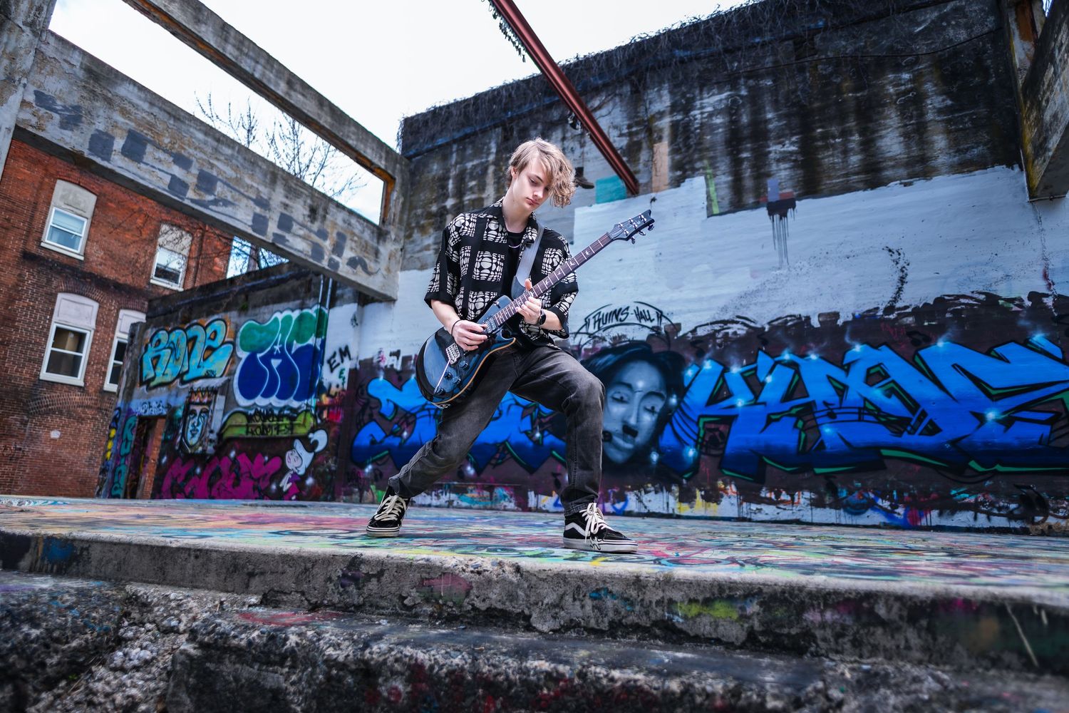 A dancer performs an energetic jump pose against a wall covered in vibrant blue graffiti art in an urban setting.