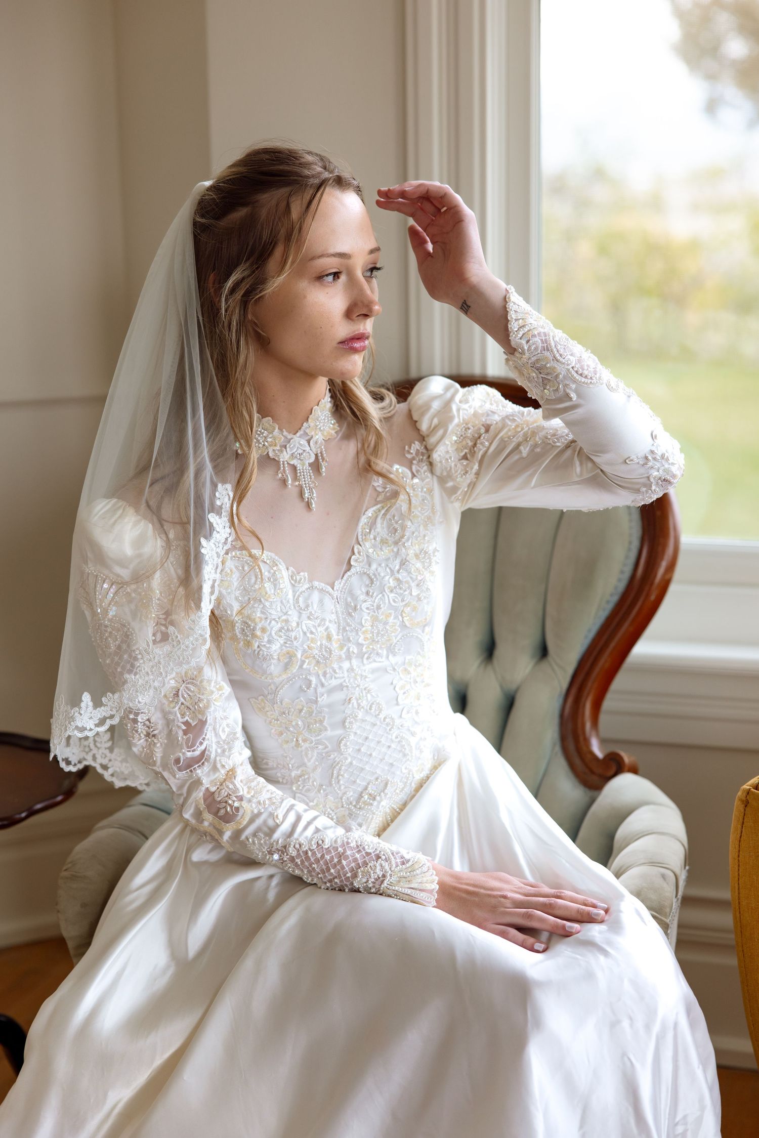 A bride in a long-sleeved lace wedding dress with delicate detailing sits gracefully by a bright window in a vintage chair.