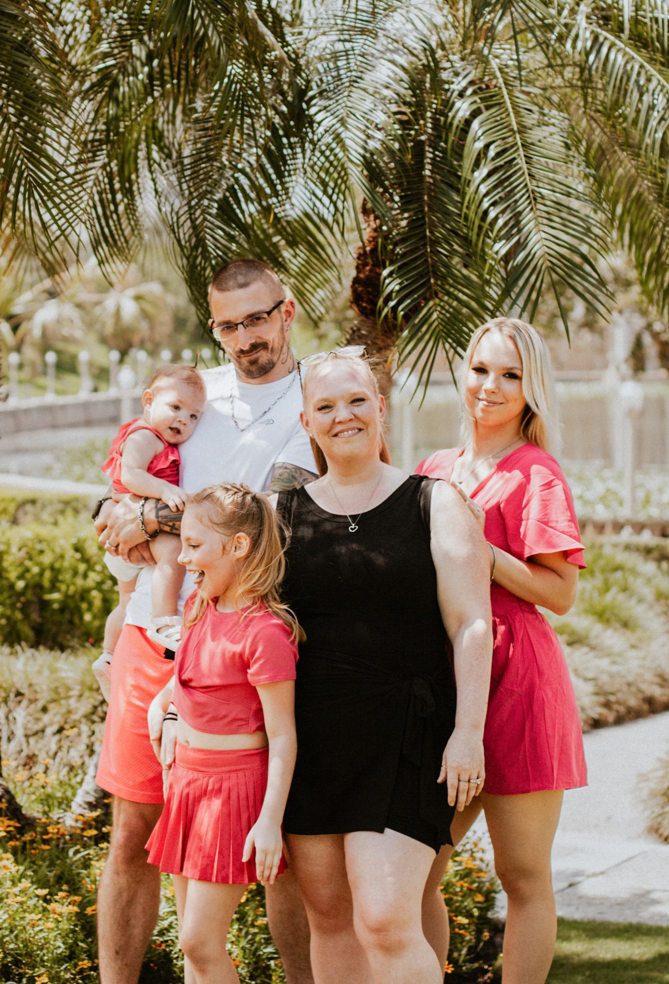 Happy family of five posing outdoors under palm trees in coordinating coral and pink outfits on a sunny day.