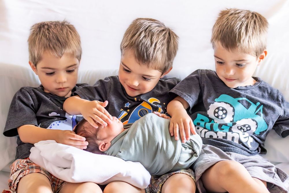 Three young boys sit together on a white couch, gently cradling and admiring a newborn baby sibling.