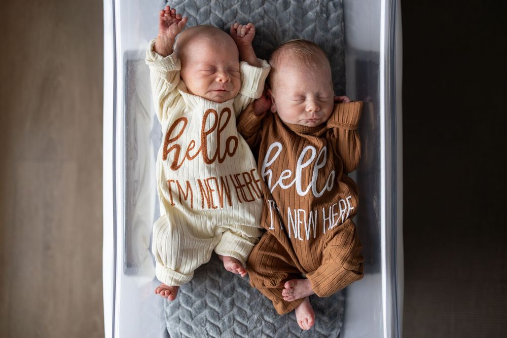 Two newborn twins sleeping side by side in a bassinet, wearing matching hello I'm new here onesies.