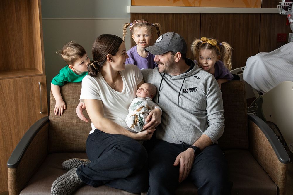 Happy family with newborn baby and three young children sitting together on a couch in a hospital room.