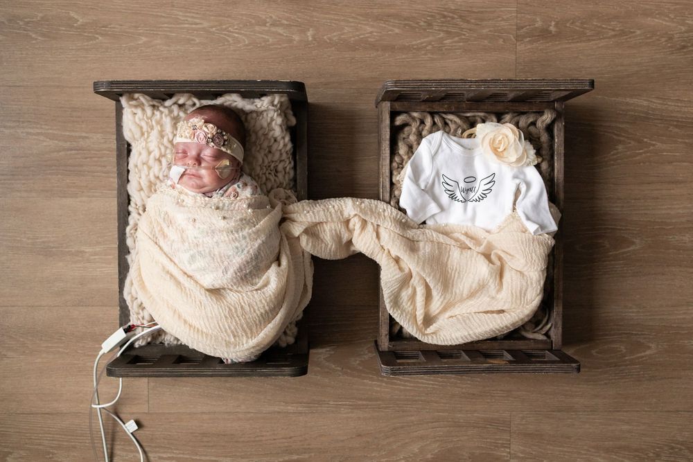 Newborn baby swaddled in cream wrap beside an angel outfit in wooden crates, twin memorial photography.