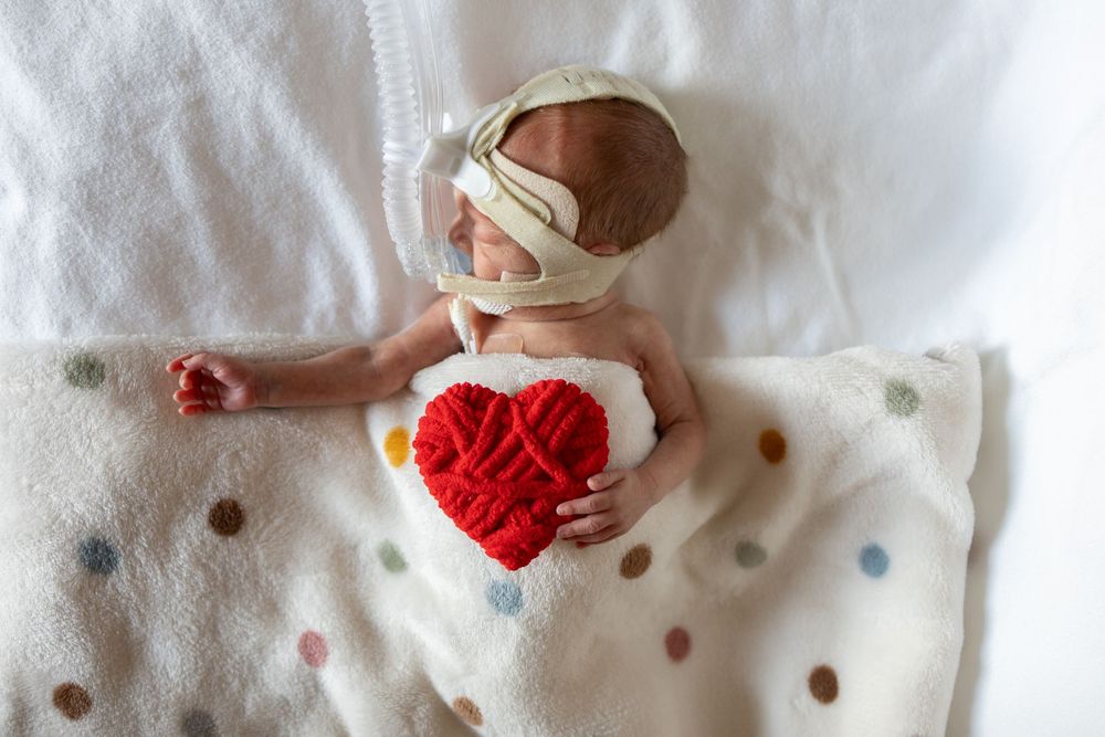 Premature newborn baby resting on polka dot blanket, wearing oxygen mask, holding a red knitted heart.