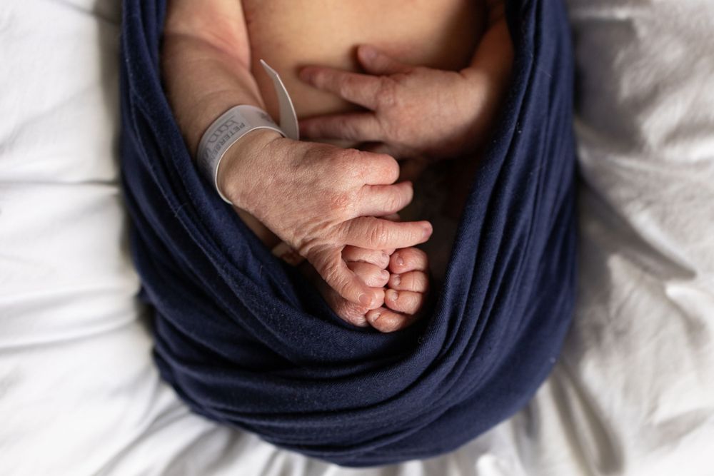 Newborn baby wrapped in navy blue swaddle blanket showing tiny hands and feet on white bedding.
