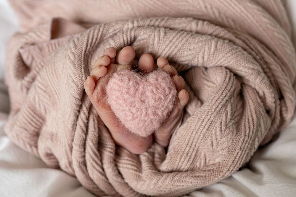 Newborn baby feet wrapped in a soft pink knit blanket with a small pink heart placed between tiny toes.