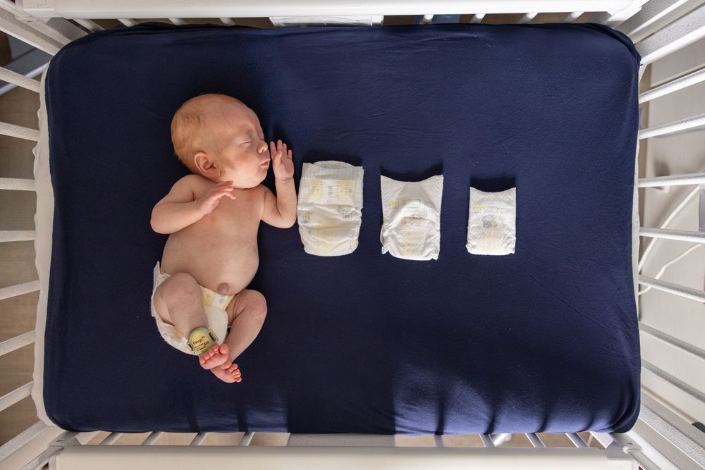 Newborn baby lying in a crib next to a row of three diapers on a navy blue sheet.