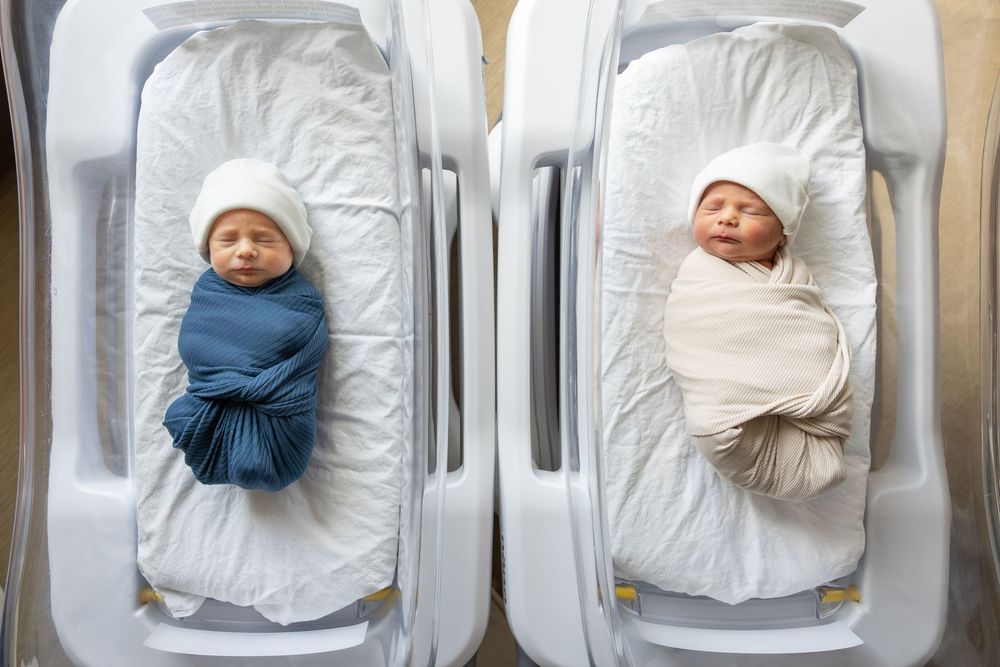 Two newborn twins swaddled in blue and white blankets lying side by side in hospital bassinets wearing white hats.