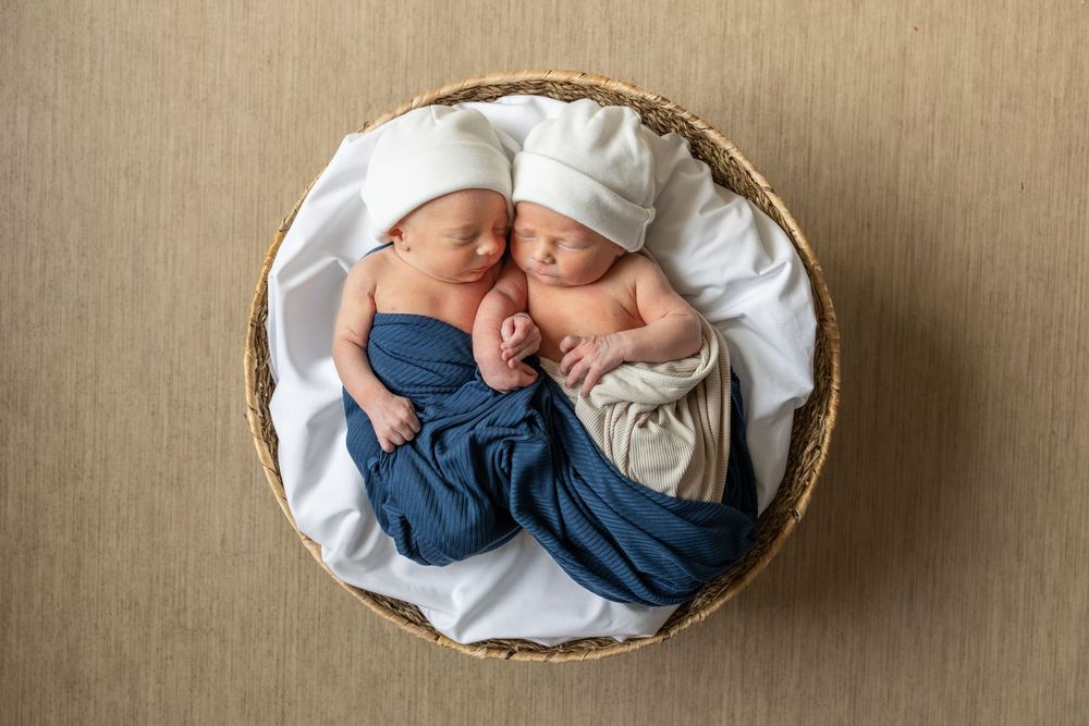Two sleeping newborn twins wrapped in blue and white swaddles, nestled together in a wicker basket.