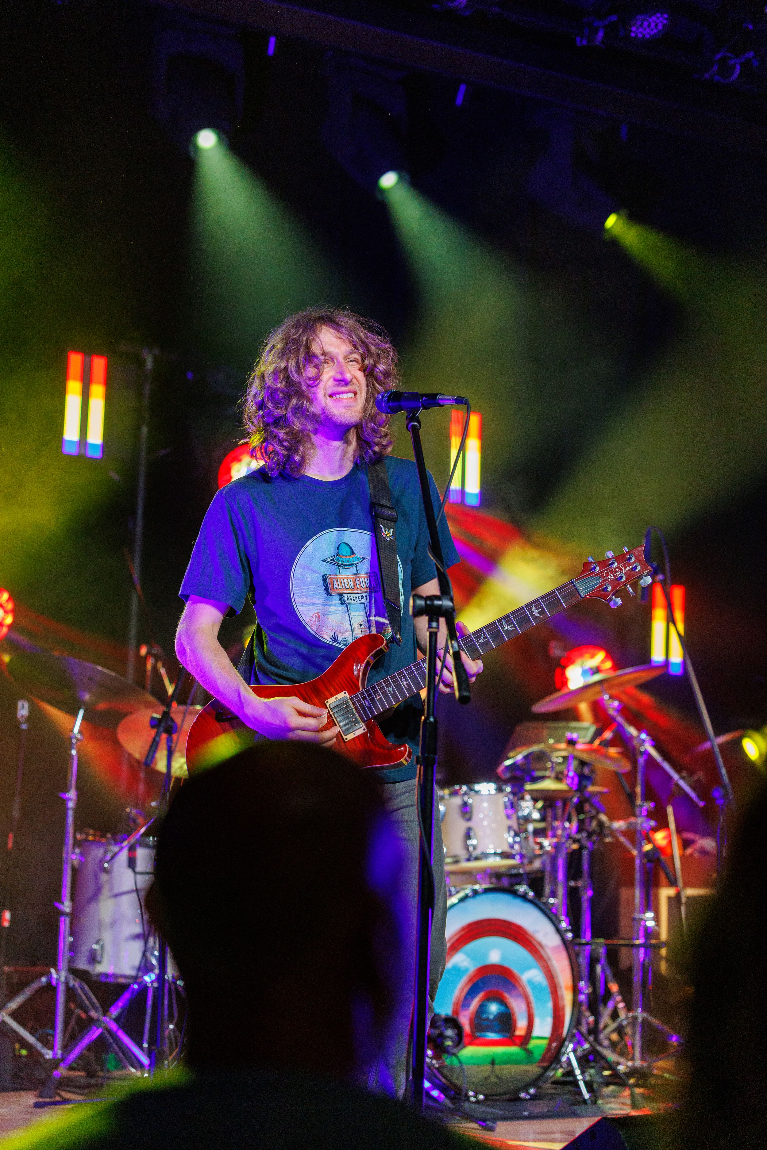 Live concert performance with musician playing electric guitar onstage with colorful lighting and drum kit in background.