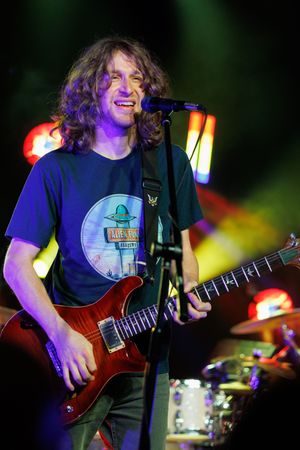 Concert performer in a blue t-shirt plays electric guitar on stage with colorful lighting effects in the background.