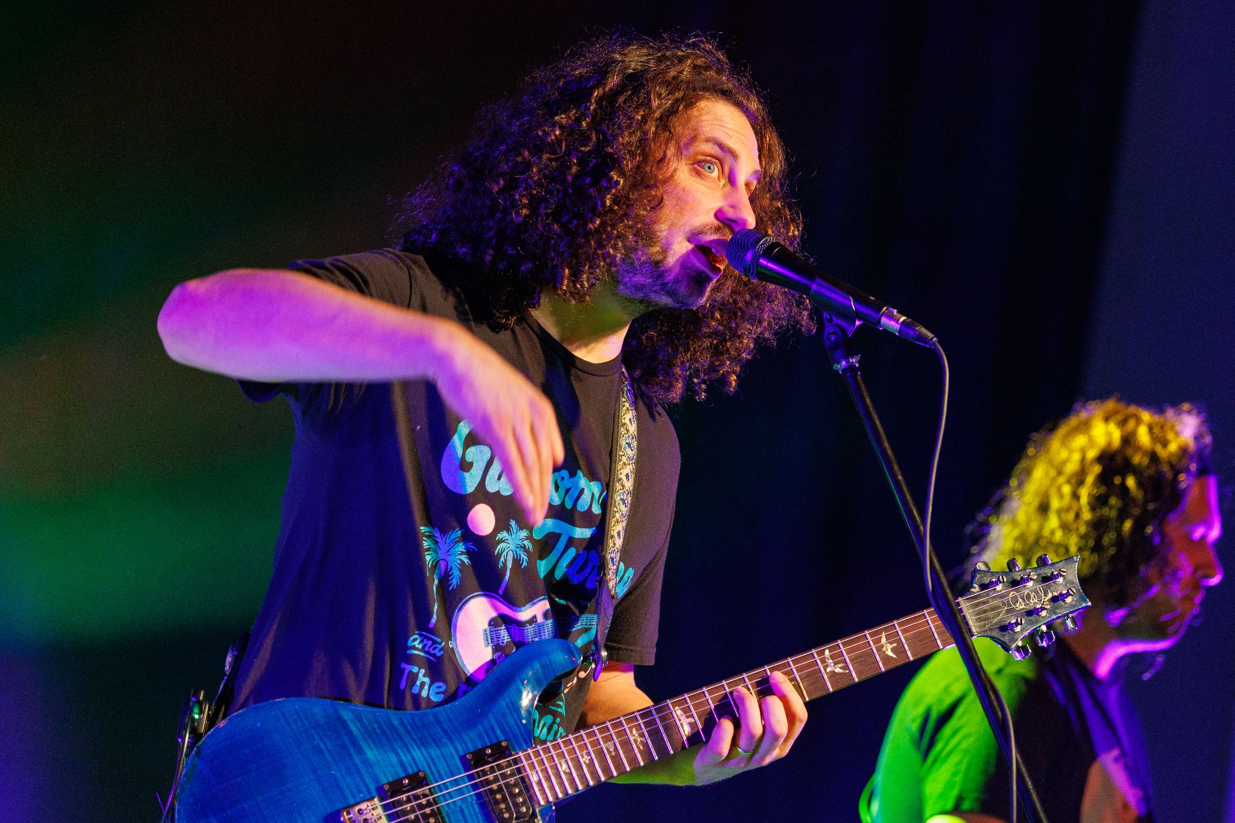 Musician with curly hair performs passionately on electric guitar with microphone on dark stage with colored lighting.