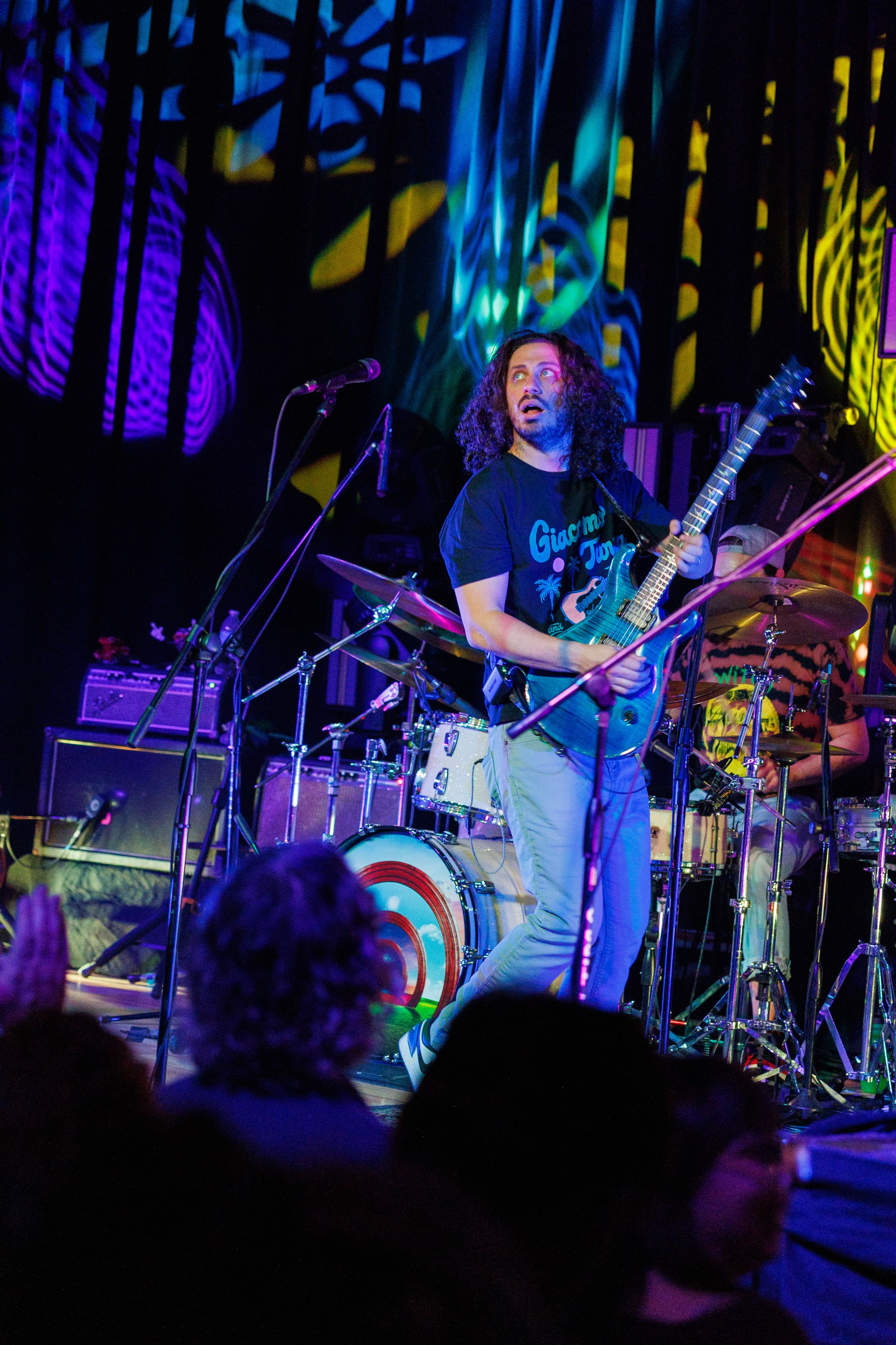 Guitarist performs on stage with colorful blue and purple lighting effects during a live concert performance.