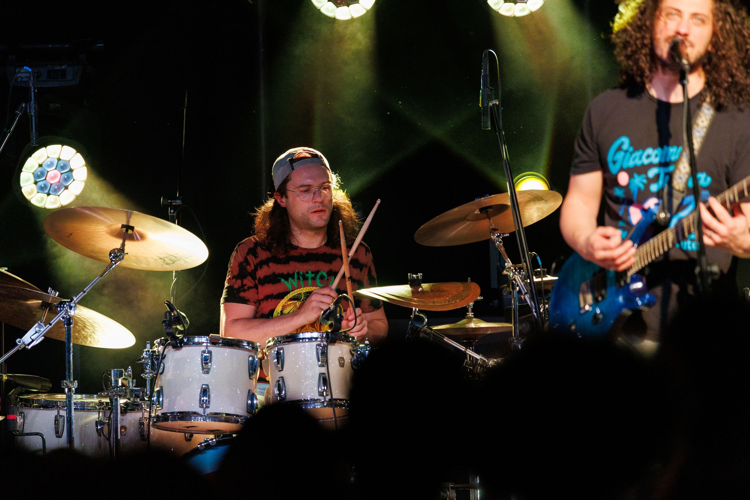 A drummer plays energetically on a drum kit while performing on stage under green stage lighting.