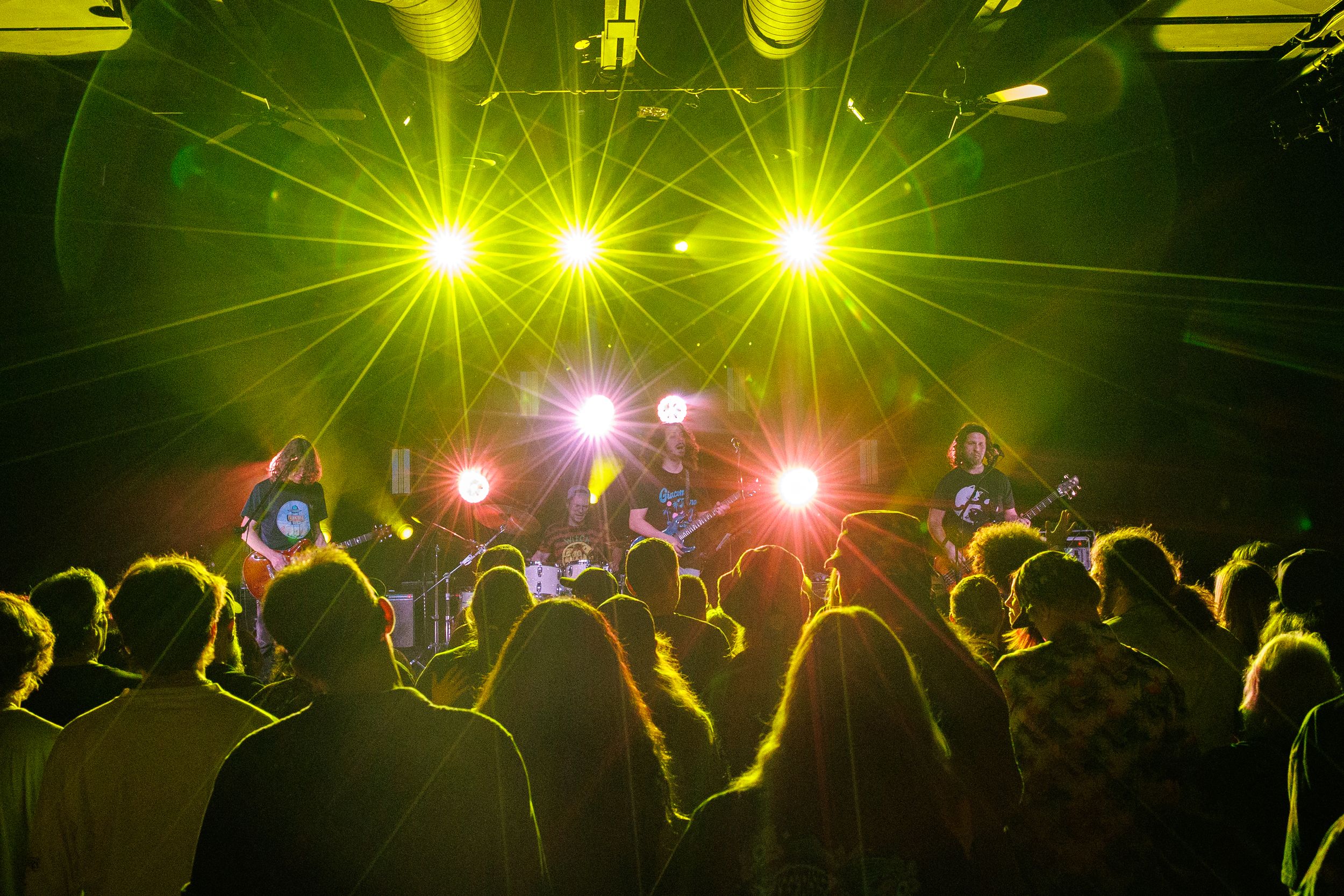Concert crowd silhouettes against bright yellow and pink stage lights during a live music performance.