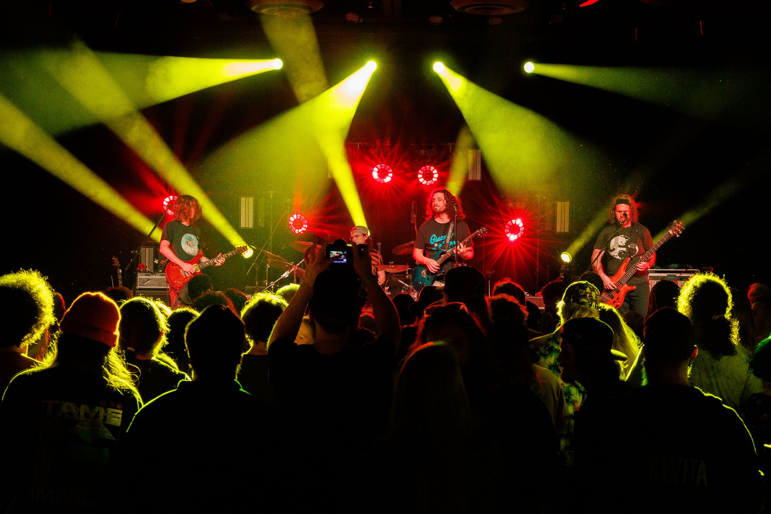 Rock band performs on stage with dramatic yellow lighting and silhouetted crowd in foreground during live concert.