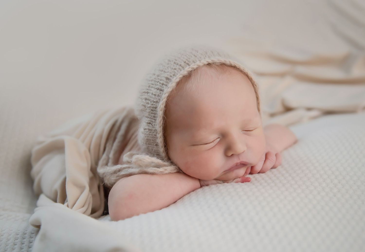 Peaceful newborn baby in beige knit wrap sleeping on white textured surface.