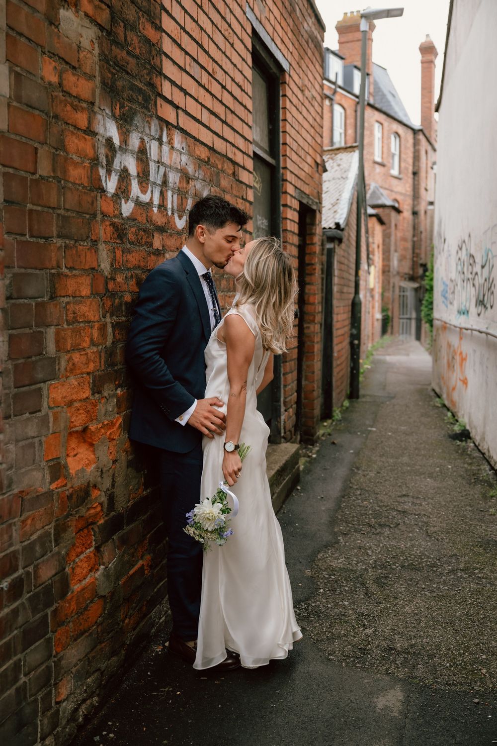 Bride and groom kissing against a brick wall in an urban alley after their elopement, bride holding a small bouquet in a white dress.