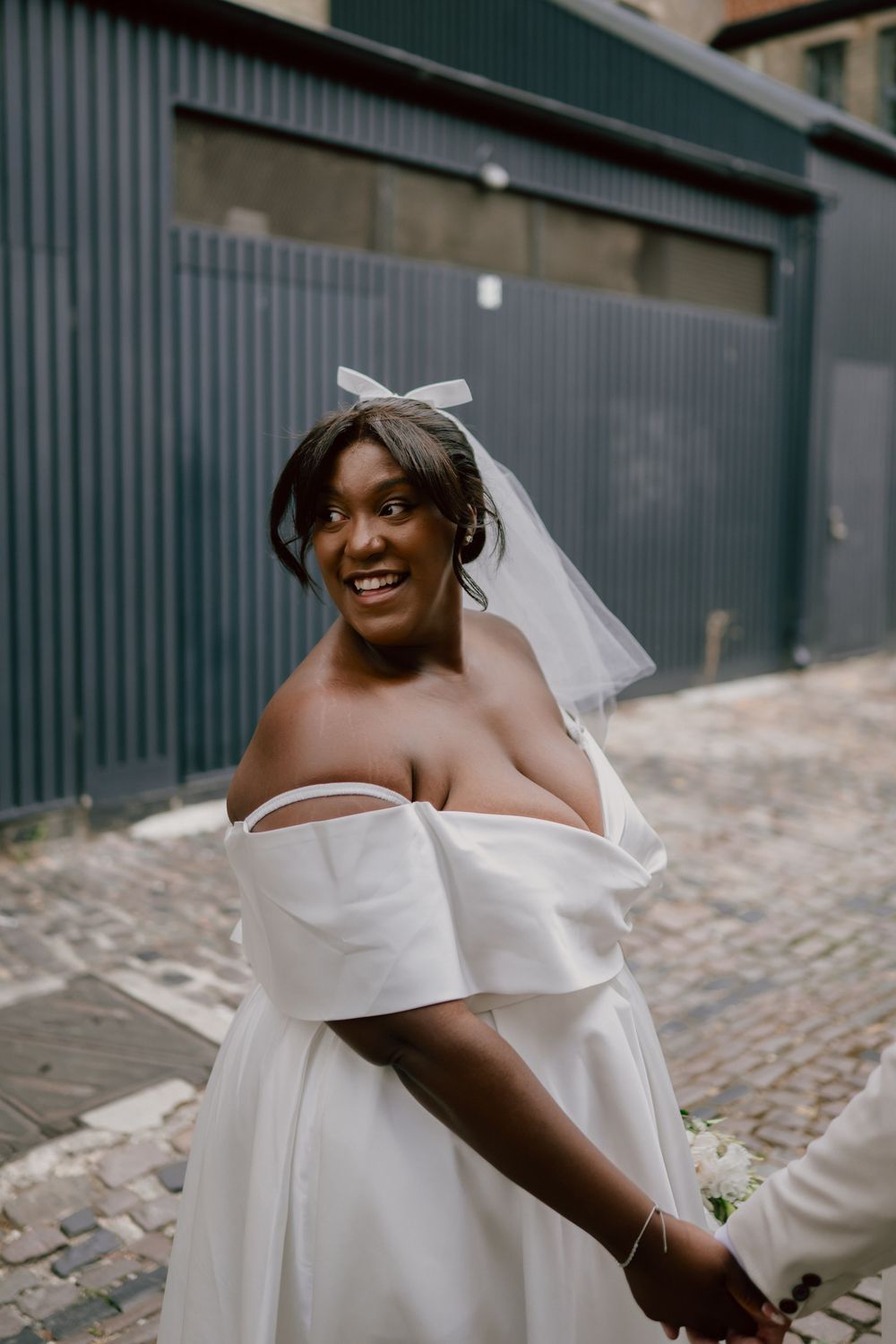 Smiling Black bride in off-shoulder white gown and veil holds hands outdoors on cobblestone street.