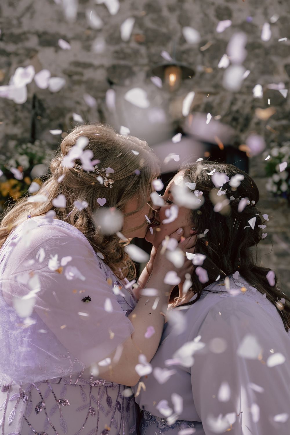 Two brides in lavender dresses share a romantic kiss surrounded by falling white heart confetti at their wedding at the Cobbles, Wales