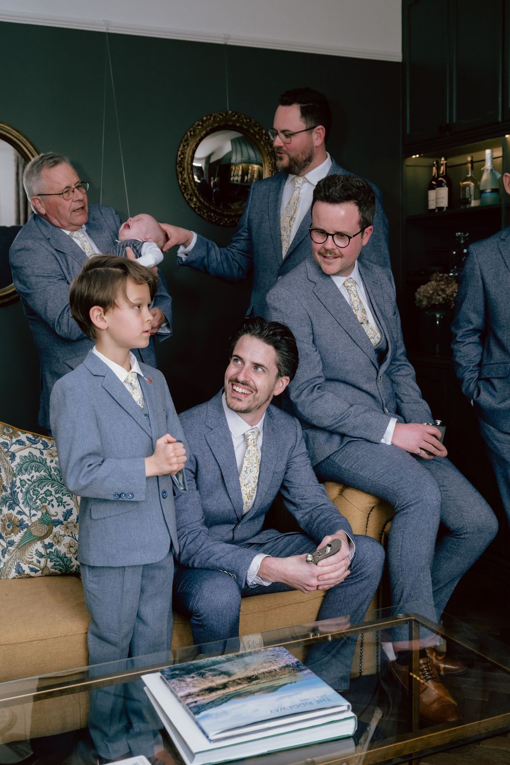 Groomsmen and ring bearer in matching grey suits with floral ties laughing together in an elegant green-walled room at the Ridgeway Barns
