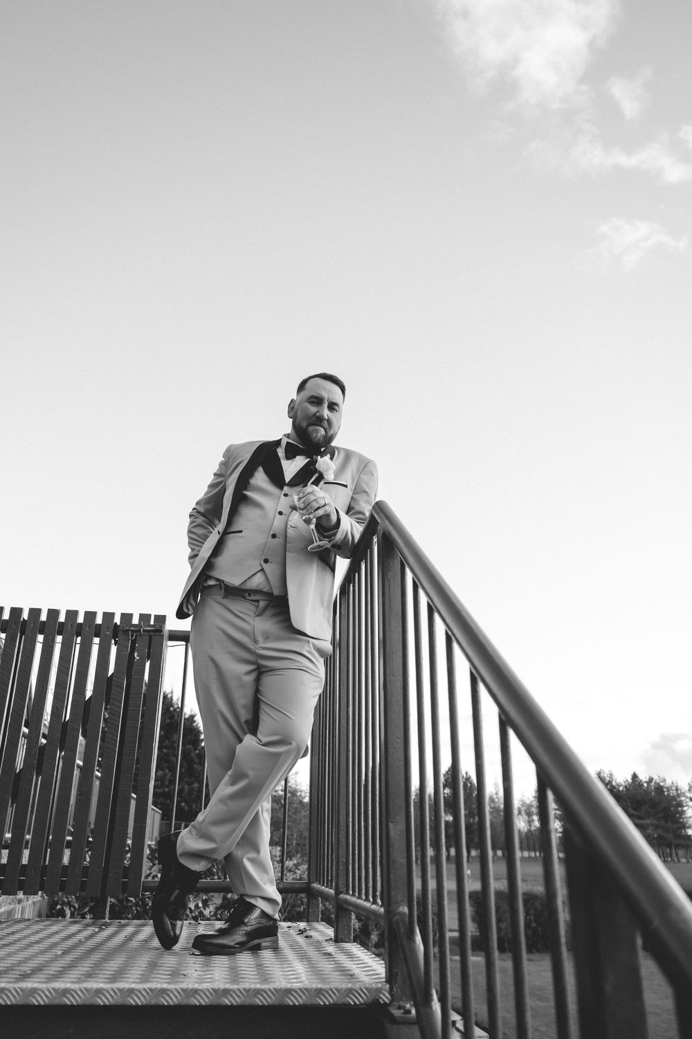 Black and white photo of a well-dressed bearded groom leaning on metal railings on outdoor steps.