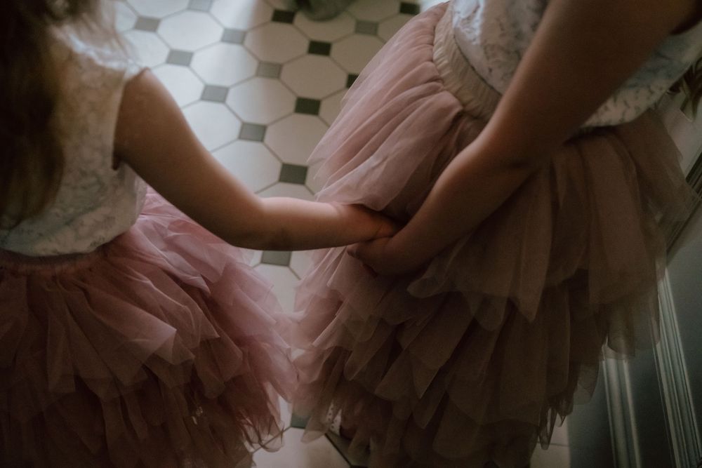 Two flower girls in matching pink tulle skirts hold hands tenderly in a softly lit room.