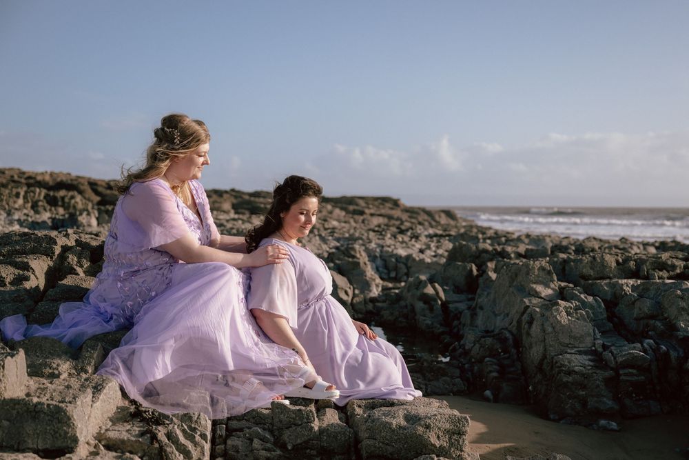Two brides in flowing lavender gowns sit on rocky coastal shore in Ogmore-by-Sea, with ocean waves in the background.