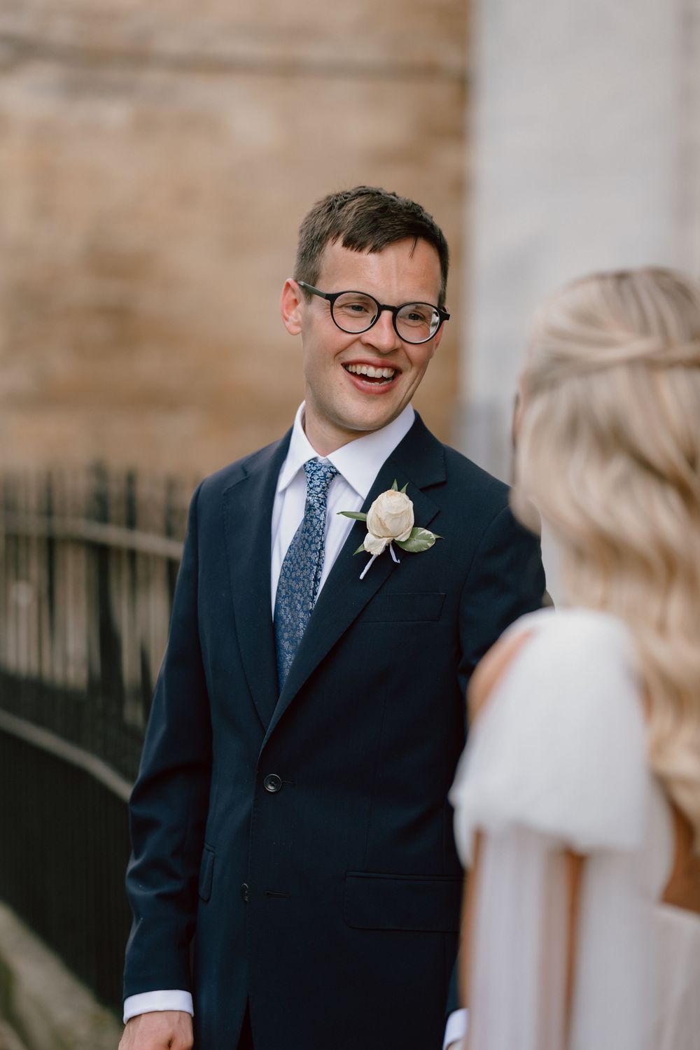 Smiling groom in navy suit with boutonniere and blue tie wearing glasses, sharing a joyful first look moment.