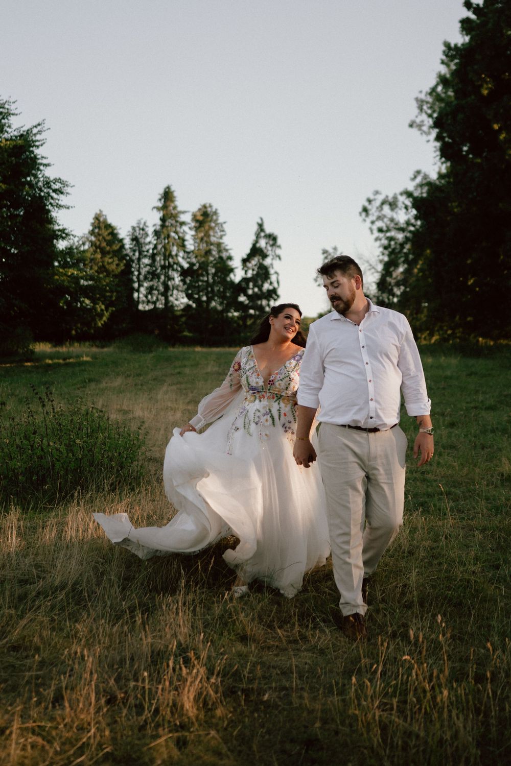 Bride and groom walk hand-in-hand through a sunlit Gloucestershire meadow, her floral whimsical wedding gown flowing in the breeze.
