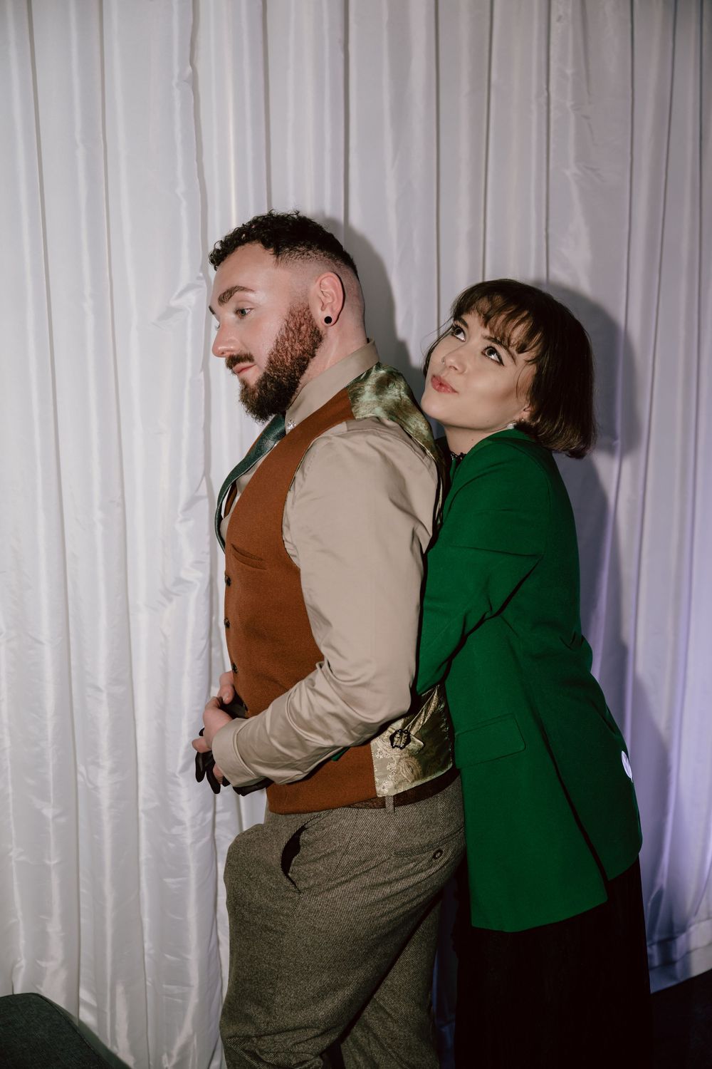 A bearded man in a brown vest and a fashion forward woman in a green blazer strike a pose at a gay wedding in Wales, in a flash photograph