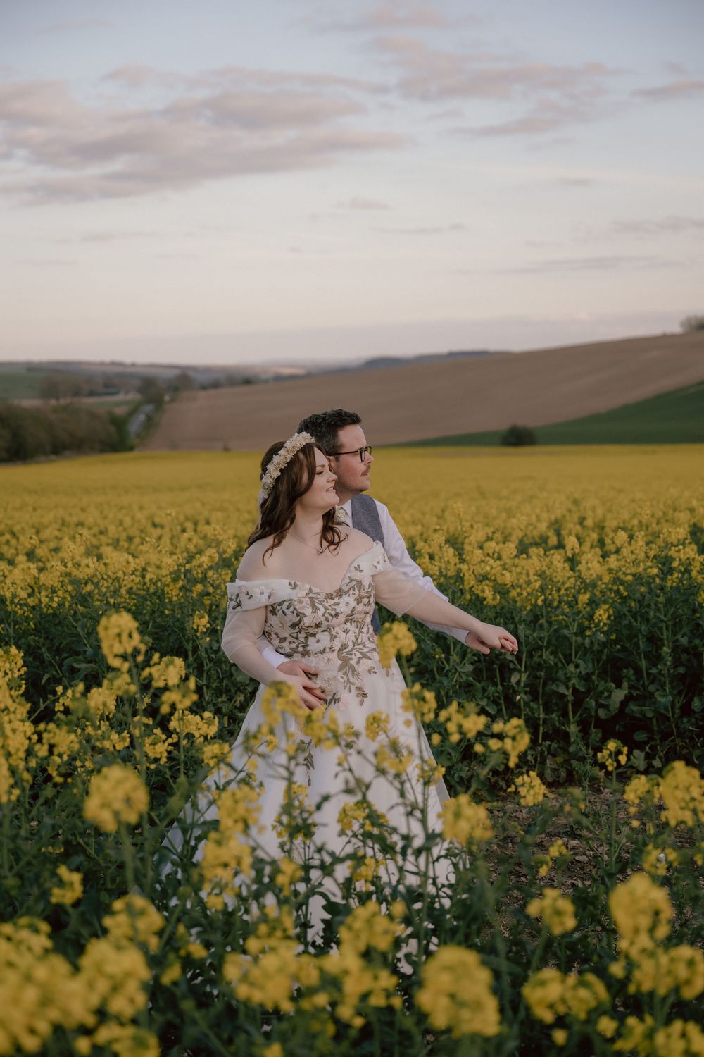 Bride and groom stand romantically in a vibrant yellow rapeseed field at sunset near the Ridgeway Barns wedding venue