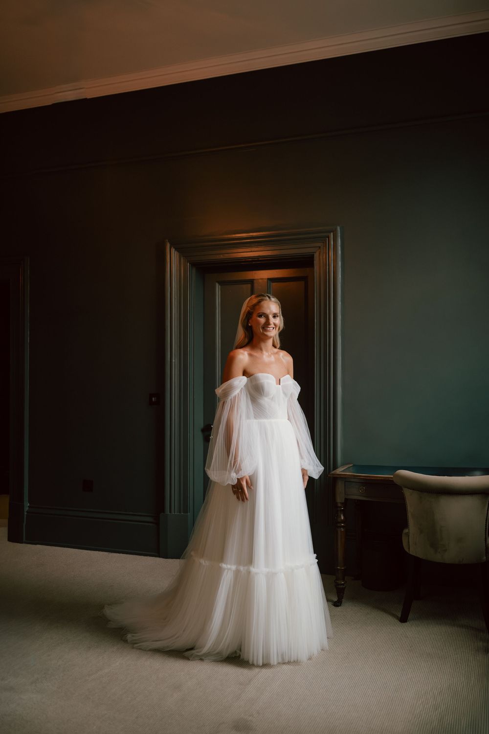 Bride in off-shoulder white tulle wedding gown standing in elegant dark teal room doorway at the Kings Head hotel in Cirencester.