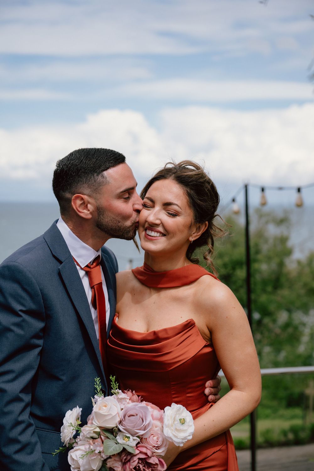 Groom in blue suit kisses bridesmaid in rust satin dress holding pink bouquet outdoors by the sea.