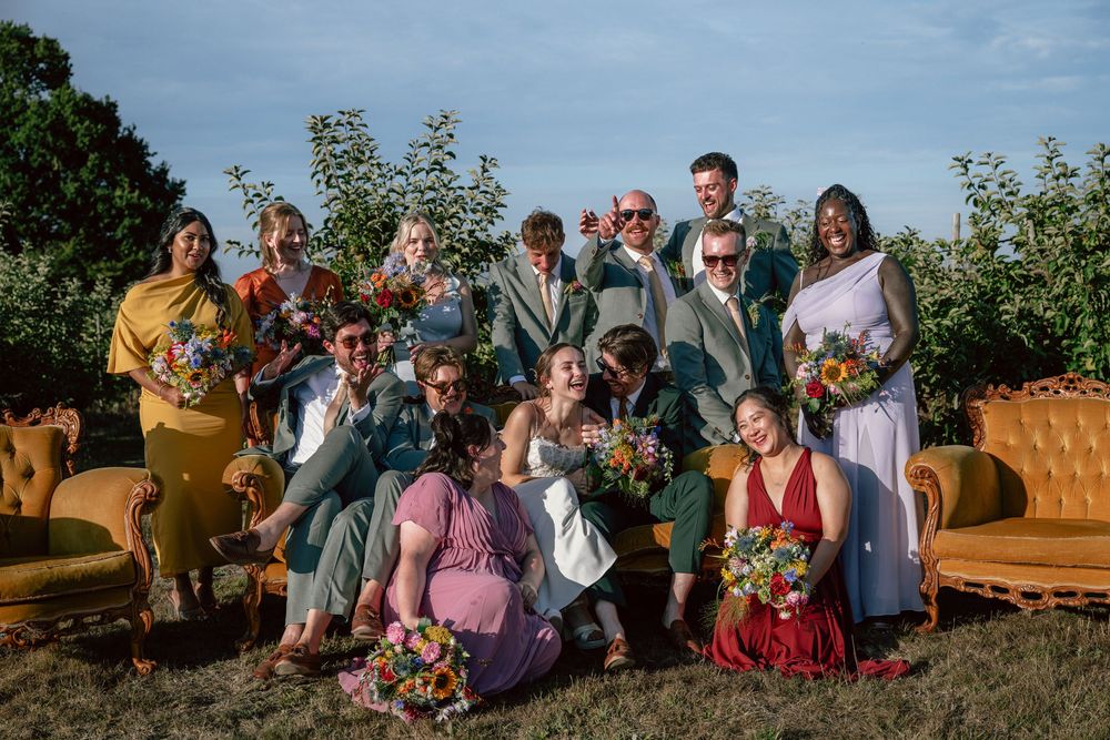 Colorful wedding party editorial posing outdoors with vintage yellow sofas, bouquets, and festive attire on a sunny day.