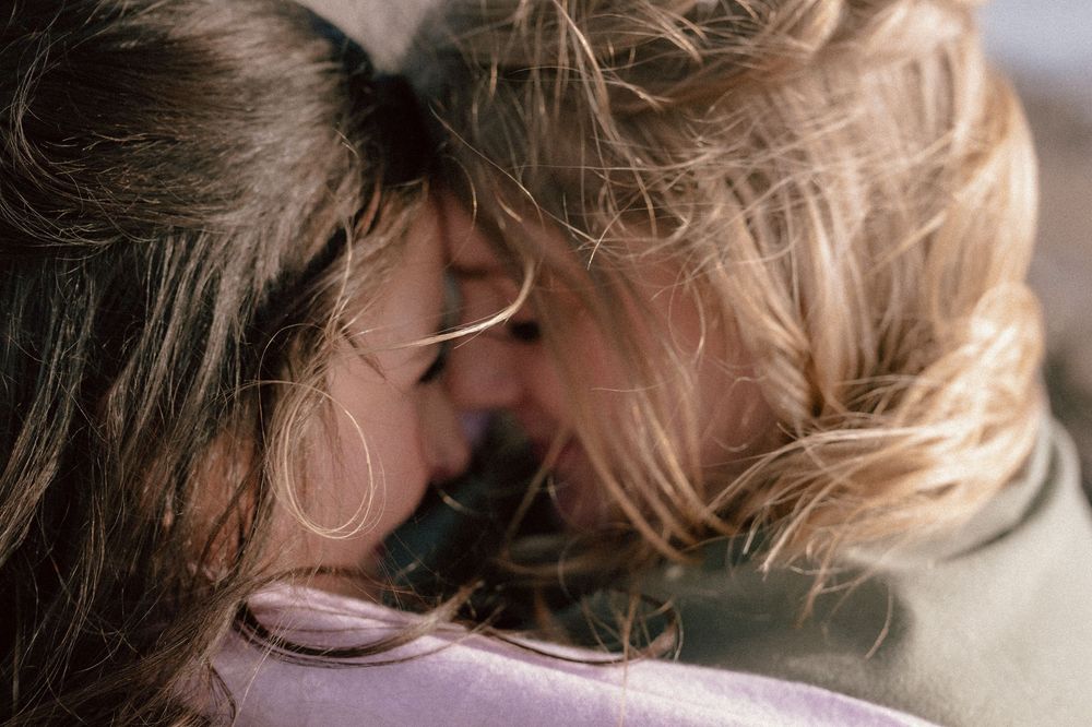 Two brides touching foreheads in a tender moment, with soft natural lighting highlighting their hair which is whipping around in the wind