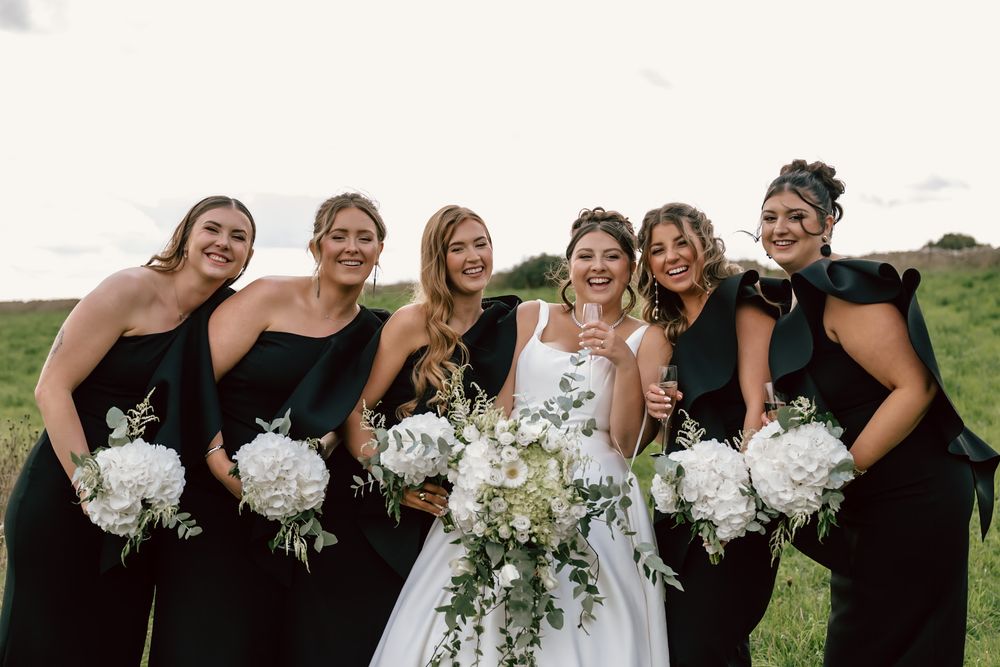 Black and white Wedding party in black dresses holds white floral bouquets while posing outdoors on a grassy hill.