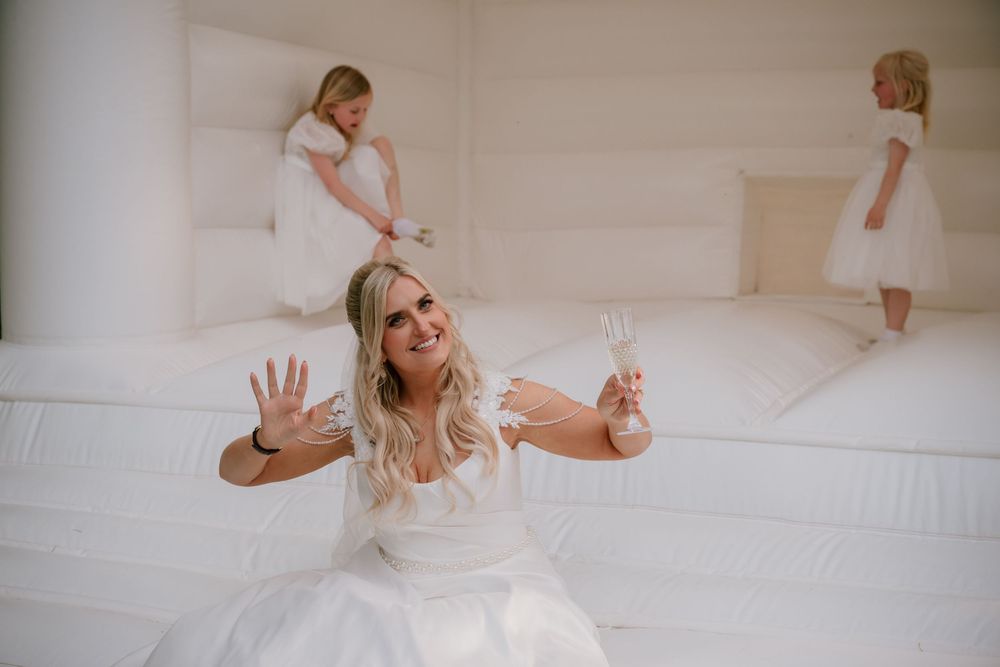 Bride sitting in a white bounce castle holding champagne, smiling at camera, with two flower girls playing behind her.