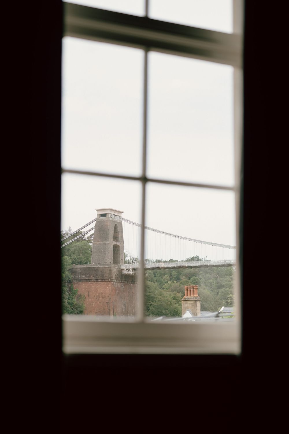 View through a multi-pane window of the Clifton Suspension Bridge tower and cables against an overcast sky.