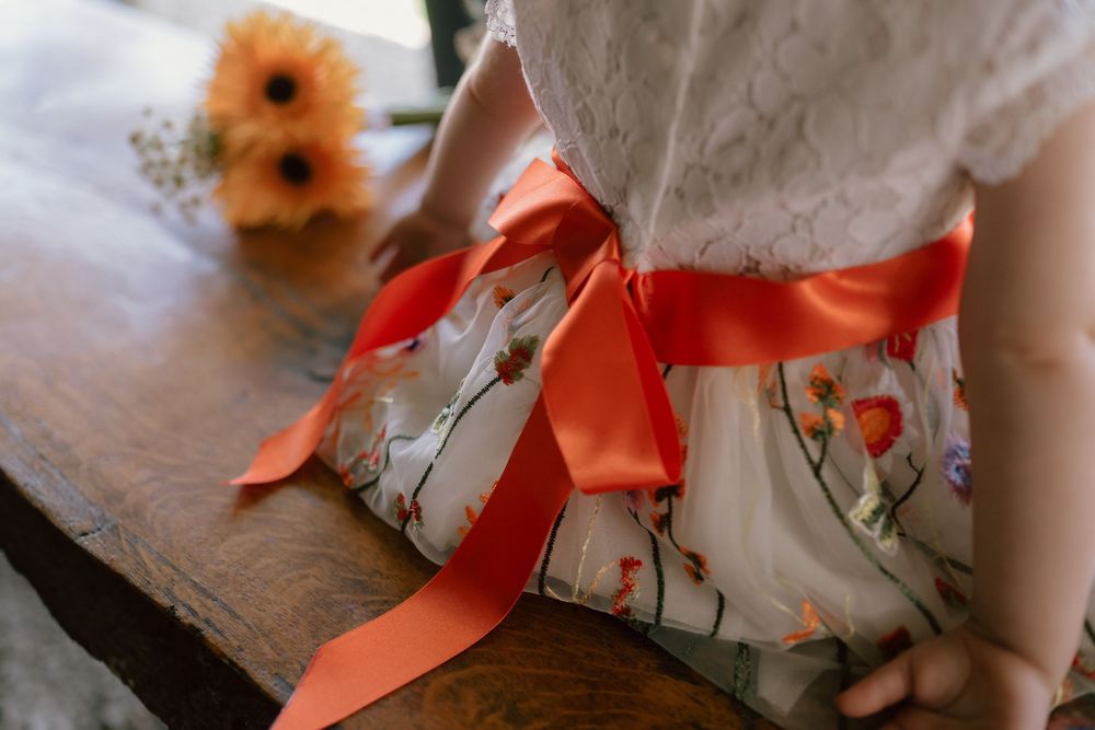 Young girl in floral dress with orange satin bow sash sits on wooden bench near sunflower bouquet.