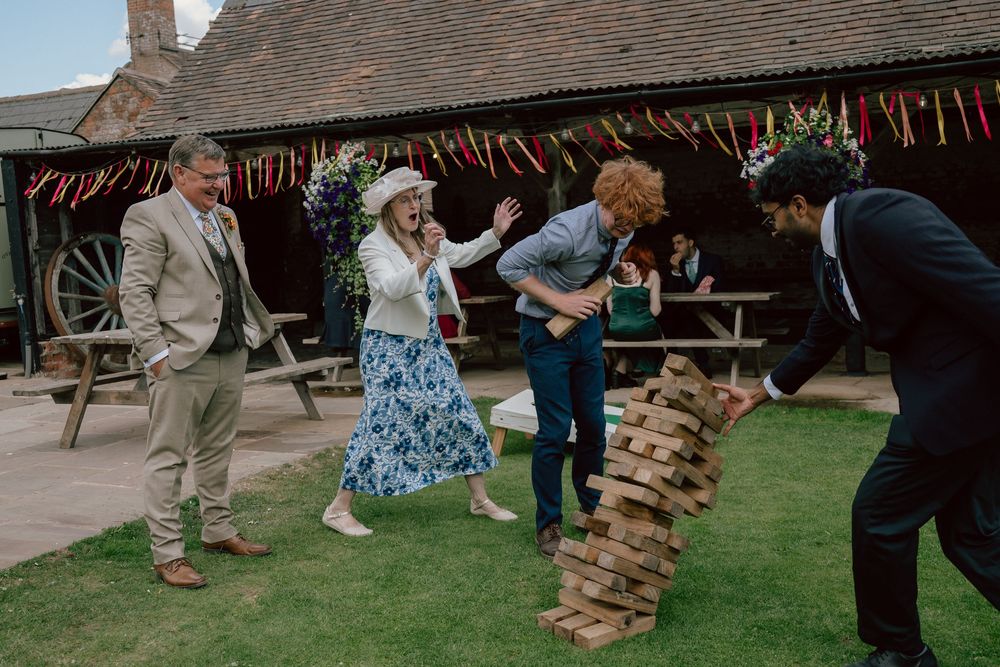 Group of friends playing giant Jenga outdoors at a rustic barn wedding venue decorated with colorful bunting.