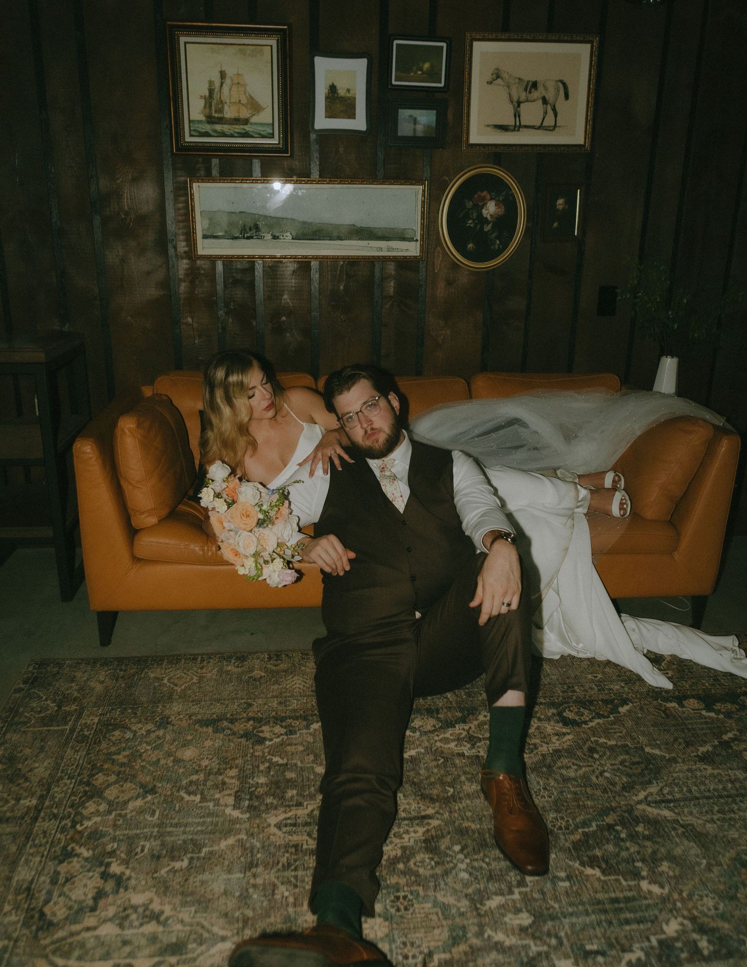Newlyweds relax on vintage orange leather couch in rustic room with framed artwork on dark wood paneled walls.