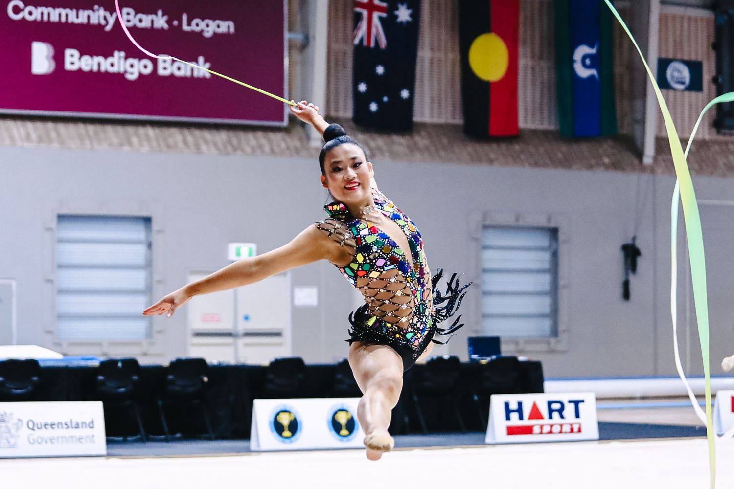 A rhythmic gymnast in a black leotard performs an elegant pose during a competition in a gymnasium decorated with flags.