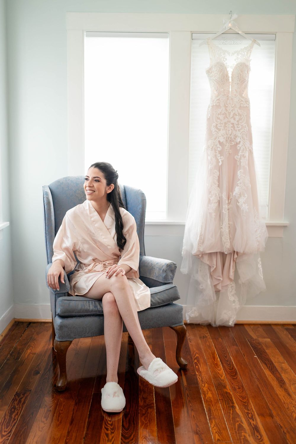 A silk robe-clad person sits in a gray armchair with slippers while a beautiful lace wedding gown hangs nearby.